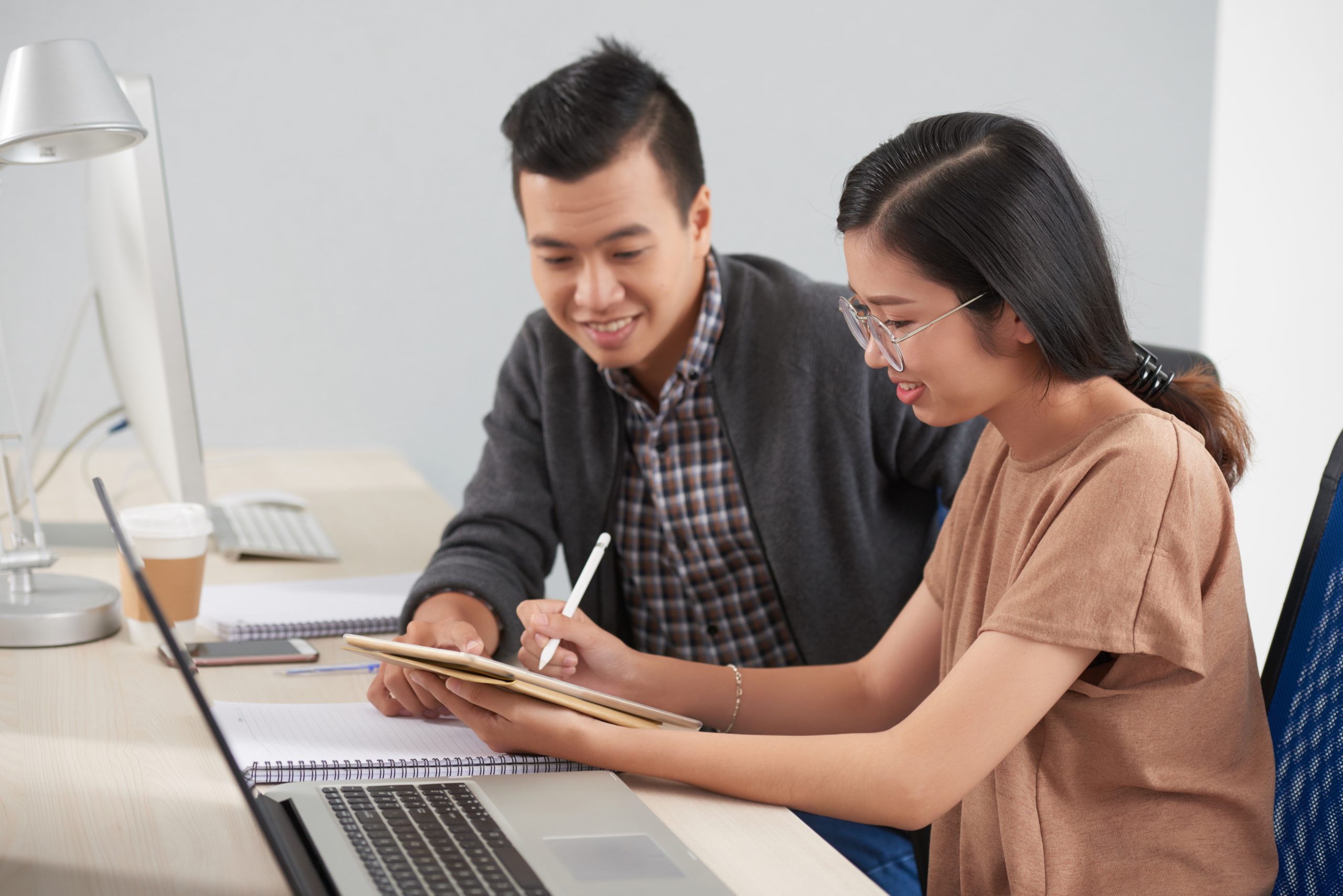 Asian young businesswoman in eyeglasses pointing at digital tablet and discussing with her colleague some working moments while they sitting at the table together at office